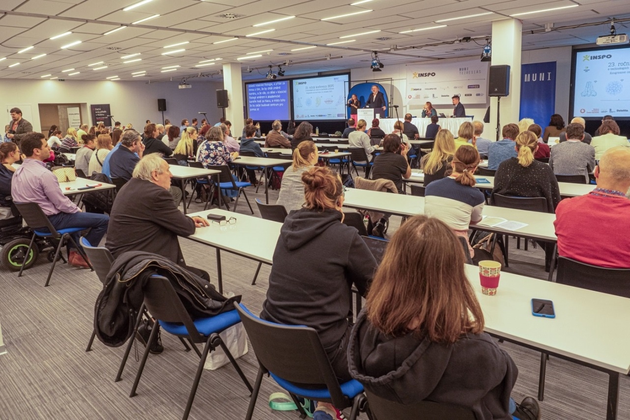 Konferenčná miestnosť plná ľudí, ktorí sedia pri dlhých stoloch. Vpredu je pódium s rečníkom.  Na stene za ním sa premietajú snímky prezentácie. Na stene sú logá rôznych spoločností a nápis INSPO a MUNI.